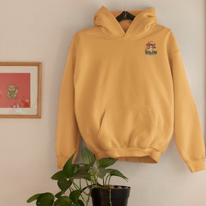 May include: A mustard yellow hoodie with a mushroom graphic on the left chest. The hoodie has a kangaroo pocket and a drawstring hood.