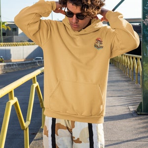 May include: A person wearing a mustard yellow hoodie with a mushroom graphic and the text "Shroom Queens". The hoodie has a kangaroo pocket and a drawstring hood. The person is wearing camouflage pants with black and white stripes.