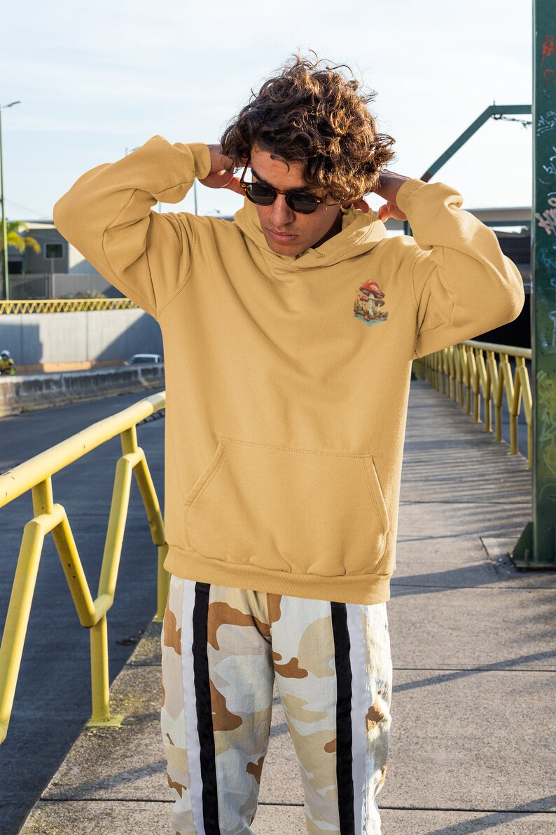 May include: A person wearing a mustard yellow hoodie with a mushroom graphic and the text "Shroom Queens". The hoodie has a kangaroo pocket and a drawstring hood. The person is wearing camouflage pants with black and white stripes.