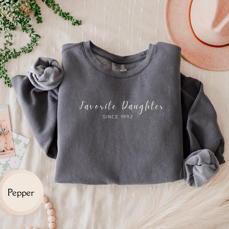 May include: A gray sweatshirt with the text "Favorite Daughter SINCE 1992" in white script. The sleeves are rolled up. The word "Pepper" is in a circle in the lower left corner.