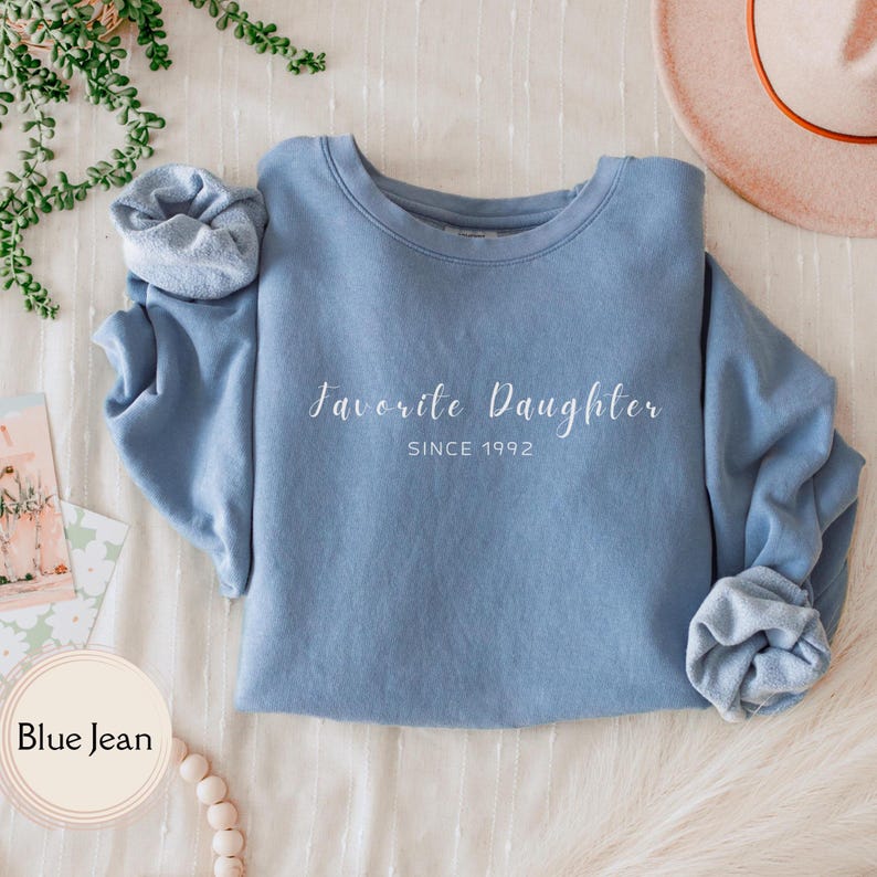 May include: A blue jean-colored sweatshirt with the words "Favorite Daughter" and "Since 1992" printed in white script. The sleeves are cuffed. The sweatshirt is displayed on a white surface.