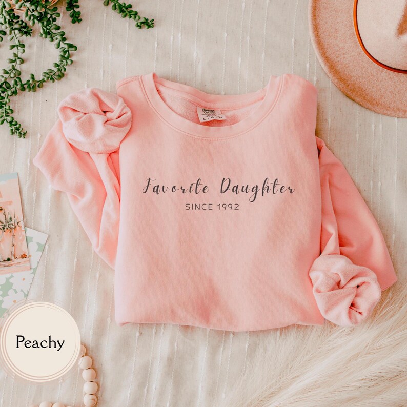 May include: A peach-colored sweatshirt with the text "Favorite Daughter SINCE 1992" in a cursive font. The long-sleeved top is folded, with the sleeves bunched up. A hat is visible in the background.