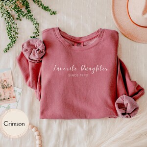 May include: Crimson-colored sweatshirt with the text "Favorite Daughter SINCE 1992" in white script. The sweatshirt is folded, with the sleeves bunched up. A hat and greenery are in the background.