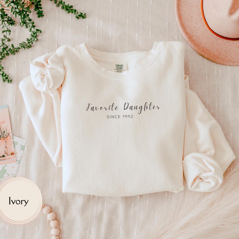 May include: Ivory-colored sweatshirt with the text "Favorite Daughter SINCE 1992" in a script font. The long-sleeved crewneck top is folded on a light-colored surface, with a hat and greenery in the background.