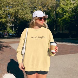 May include: A butter-colored sweatshirt with the text "Favorite Daughter Since 1992" in a script font. The sweatshirt is worn with black shorts, a white cap, and sunglasses. A person is holding a coffee cup.