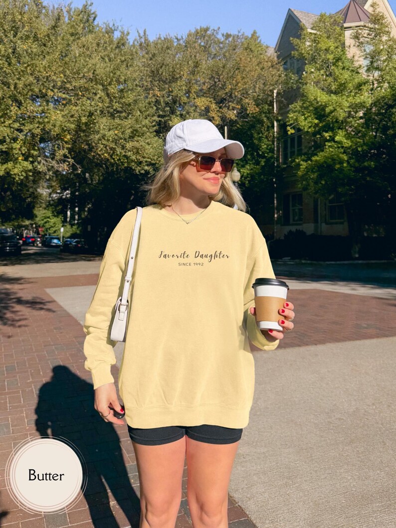 May include: A butter-colored sweatshirt with the text "Favorite Daughter Since 1992" in a script font. The sweatshirt is worn with black shorts, a white cap, and sunglasses. A person is holding a coffee cup.