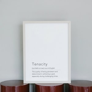May include: A white framed print with the word "Tenacity" and its definition in black text. The print is sitting on three brown cylindrical objects.