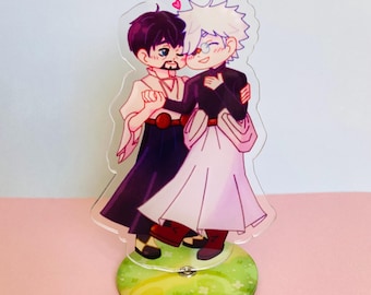 Puede incluir: Un expositor de acrílico con dos personajes de estilo anime, uno con cabello oscuro y barba y el otro con cabello blanco, ambos con ropa fluida y tomados de la mano. Un pequeño corazón rosa se sitúa sobre sus cabezas.