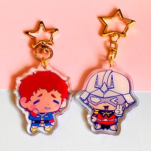 May include: Two acrylic keychains featuring cartoon characters with gold-colored star-shaped clasps. The left keychain depicts a character with red hair in a blue and red uniform. The right keychain shows a character in a white helmet and red suit.