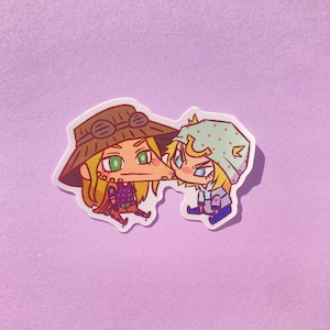 May include: A sticker featuring two cartoon characters. One character wears a brown hat with goggles and a purple outfit. The other character wears a light blue hat with pink polka dots and a gray outfit. The sticker has a white border and is on a purple background.