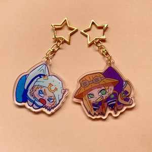 May include: Two acrylic keychains featuring cartoon characters. One keychain depicts a character in a blue outfit, the other in a brown hat and purple outfit. Both keychains have gold star-shaped clasps.