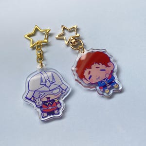 以下が含まれることがあります： 漫画のキャラクターデザインのアクリルキーホルダー2つ。1つのキーホルダーは、白いヘルメットと赤と黒の衣装を着て、ピースサインをしているキャラクターが特徴です。もう1つのキーホルダーは、赤毛のキャラクターが青と赤の衣装を着ています。どちらも金色の星型の留め金が付いています。