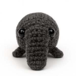 May include: A handmade, dark gray crocheted creature with large, glossy black eyes. The amigurumi-style plush has a rounded body, a trunk-like snout, and small feet. The toy is set against a plain white background.