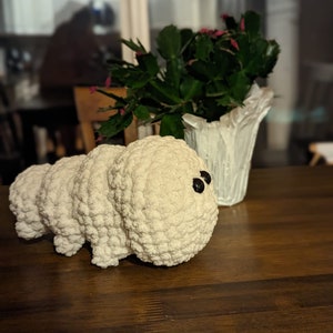 May include: A white crocheted stuffed animal shaped like a caterpillar with black eyes. The caterpillar is sitting on a wooden table next to a potted plant.