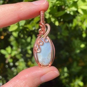 May include: A handcrafted copper wire pendant with a light blue gemstone. The teardrop-shaped stone is encased in intricate copper wire, with decorative swirls at the top. The pendant is held up by a finger against a blurred green background.
