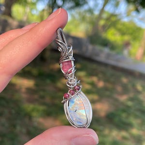 May include: A handcrafted pendant featuring a large, iridescent moonstone, accented with pink tourmaline and small red gemstones. The stones are intricately wrapped in silver wire, creating a unique and elegant design. The pendant is held in a hand.