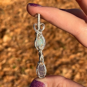 May include: A silver wire-wrapped pendant featuring a clear iridescent opal and a purple amethyst. The pendant is held by a finger with purple glitter nail polish.