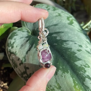 May include: A handcrafted pendant featuring a large, polished, pink gemstone, accented with smaller white and red gemstones. The stones are intricately wrapped in silver-colored wire, creating a unique and artistic design. The pendant is suspended from a silver bail.