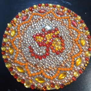 Peut inclure: Une pièce ronde décorative en matériau brun avec une mosaïque de petits strass colorés. Les strass sont disposés en cercle avec un motif central de strass rouges, orange et jaunes.