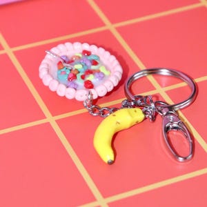 May include: A novelty keychain featuring a miniature bowl of cereal with a spoon and a yellow banana charm. The cereal bowl is pink with colorful cereal pieces. The keychain has a silver clasp and ring.
