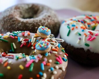 May include: Close-up of three donuts with colorful sprinkles. One donut is covered in chocolate frosting, another in white frosting, and the third is dusted with a light brown coating. A donut-shaped keychain with blue frosting and sprinkles is in the foreground.