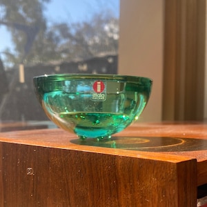 May include: A small, translucent emerald green glass bowl with a rounded shape and a smooth finish. A red and white sticker on the front displays the text iittala FINLAND. The bowl features a thick base and is crafted from clear green glass.