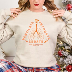 Könnte beinhalten: Ein beiger Pullover mit einer orangefarbenen Grafik von einer Lunge mit den Worten "Ventilate, Intubate, Sedate" darum herum. Der Text unter der Grafik lautet "Beside You Breath by Breath, Beat by Beat."
