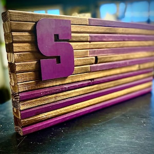 May include: Wooden wall art featuring a layered design with alternating gold and purple planks. A large purple number "5" is affixed to the front. The piece is constructed from wood and has a rustic, handcrafted appearance.