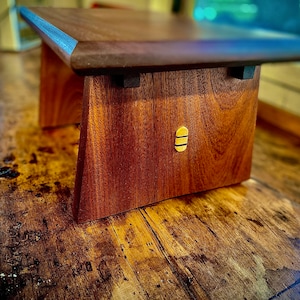 May include: A handcrafted wooden stool with a dark brown top and reddish-brown legs. The stool features a unique design with a small, gold-colored accent on the front. The stool is made of solid wood and appears to be sturdy.