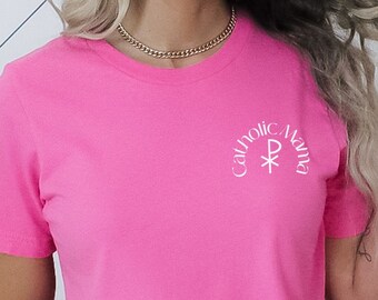 Peut inclure: Un t-shirt rose avec un motif blanc qui dit "Catholic Aura" avec une croix stylisée.