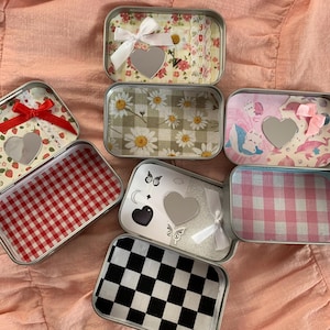 Könnte beinhalten: Eine Sammlung rechteckiger Metalldosen mit verschiedenen dekorativen Designs. Einige Dosen weisen florale Muster, Vichy-Karo oder herzförmige Spiegel auf. Andere haben Schmetterlings- und Schleifenakzente, mit Farben wie Rot, Rosa und Schwarz.