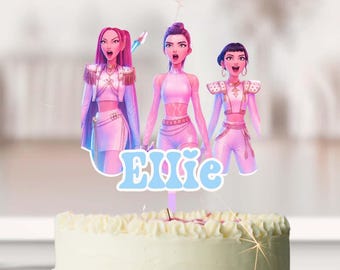 May include: A round cake with a white and pink ombre design, topped with a cake topper featuring three cartoon figures and the name "Ellie" in blue. The cake sits on a white cake stand.