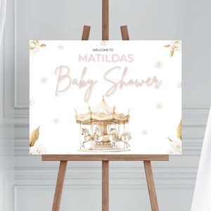 Może przedstawiać: Znak baby shower na drewnianym sztaludze. Znak jest biały ze złotymi i różowymi akcentami. Zawiera ilustrację karuzeli i słowa "Welcome to Matildas Baby Shower."