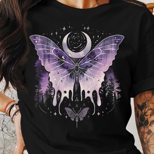 May include: Black t-shirt featuring a large, detailed butterfly design. The butterfly has purple and white wings with a crescent moon and stars in the background. A smaller butterfly is below the main design, with a forest scene in the background.