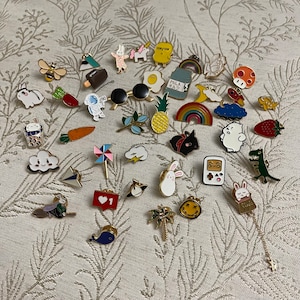 Puede incluir: Una colección de 30 pines de esmalte coloridos con varios diseños, incluyendo animales, comida y objetos cotidianos. Los pines están dispuestos sobre un fondo de tela estampada.