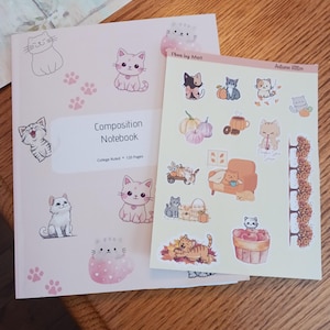 May include: A composition notebook and sticker sheet featuring cute cat illustrations. The notebook has a pink cover with cat drawings and paw prints. The sticker sheet has autumn-themed cat designs, pumpkins, and a coffee cup.