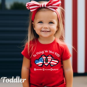 Może przedstawiać: Uśmiechnięte małe dziecko w czerwonej koszulce z napisem "On Fridays We Wear RED" i "Remember Everyone Deployed". Koszulka ma trzy serca z motywem amerykańskiej flagi. Dziecko ma r&oacute;wnież czerwoną opaskę na głowie z kokardą.