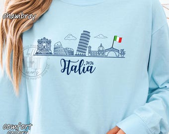 May include: Light blue long-sleeve sweatshirt with a graphic of Italian landmarks in navy blue. The word "Italia" is written in cursive, with "2026" below it. The sweatshirt also has the words "comfort colors unisex".