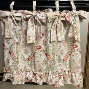 May include: A floral patterned kitchen curtain with a patchwork design. The curtain features a repeating pattern of red roses, green leaves, and beige squares. The top has decorative bows, and the bottom has a ruffled edge.