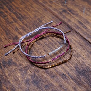May include: Four handmade bracelets with adjustable cord closures. The bracelets are in shades of purple, brown, and light blue, with small gemstone beads. The bracelets are displayed on a wooden surface.