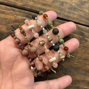 以下が含まれることがあります： 茶色、ピンク、緑、黒の色合いの様々な天然石ビーズを使用した5つのビーズブレスレット。 ブレスレットは濃い色のコードに通されており、丸、楕円、不規則なビーズなど、さまざまな質感と形状が特徴です。