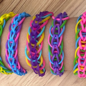 May include: Five colorful rubber band bracelets in yellow, blue, purple, green, and pink. The bracelets are stacked on a person's arm.