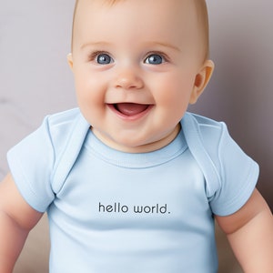 May include: A baby wearing a light blue bodysuit with the text "hello world." printed on the front. The baby is sitting on a light brown surface and smiling.