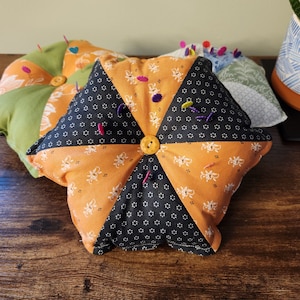 May include: Three fabric pincushions in different shapes and patterns. The largest pincushion is triangular and features orange, black, and white fabric with a button in the center. The other two pincushions are round and feature green and white fabric.