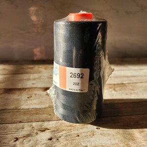 May include: A spool of black thread with a red top, wrapped in clear plastic. The label reads "2692 2DZ MADE IN ITALY". The thread is on a wooden surface.