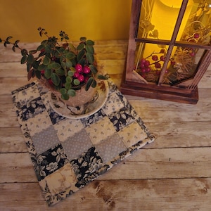 May include: A square quilted placemat with a patchwork design of floral and solid fabric squares in cream, navy, and light blue. A small potted plant with red berries sits on a saucer atop the placemat. A wooden lantern with a lit candle is in the background.