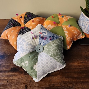 May include: Three handmade pin cushions in a starburst design. The cushions feature a variety of floral and geometric patterned fabrics in orange, green, blue, and white. Each cushion has a button center and is adorned with colorful pins.