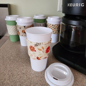 May include: Five white coffee cups with different patterned fabric sleeves. One cup has a floral pattern, another has a green and white geometric pattern, and two have a brown and white pattern. One cup has a lid. The cups are in front of a black Keurig coffee maker.