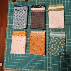 May include: Six stacks of folded fabric in various patterns and colors, including floral, geometric, and solid designs. The fabrics are arranged on a green cutting mat, showcasing a variety of quilting materials.