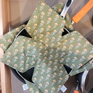 May include: A star-shaped fabric pan protector in olive green with a repeating pattern of small yellow flowers and white stems. The quilted textile features stitched lines and is designed to sit between stacked cookware to prevent scratching.